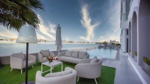 Zona relax esterna moderna con piscina, arredi bianchi e vista sull'acqua e sullo skyline della città.