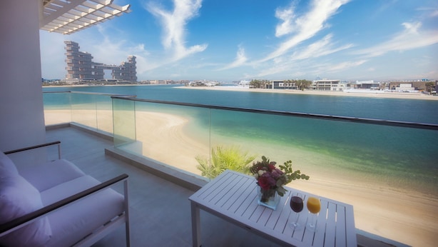 Un balcone con vista su una spiaggia e un porto turistico, con un tavolo bianco, un vaso di fiori e due bicchieri di bevande.