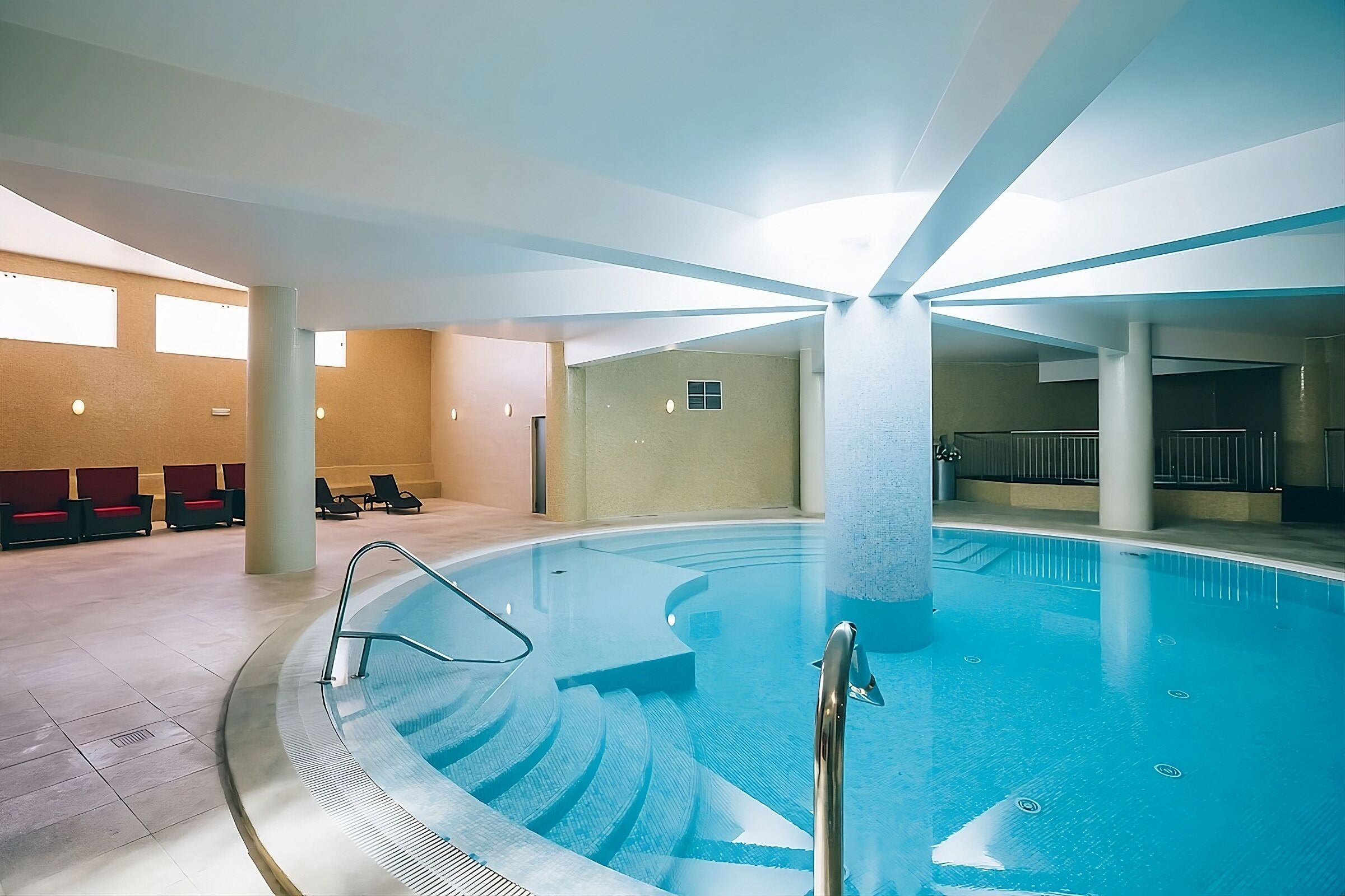 Una piscina interior con agua cristalina, una escalera central y zonas de estar con sillas rojas y negras.