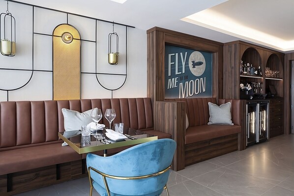 Un interior moderno con un sofá de cuero marrón, una mesa de vidrio y una silla azul. Se observa una pared con el letrero 'FLY ME TO THE MOON' y un mueble de madera con cristalería.