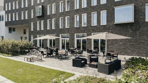 A modern building with a brick facade, outdoor seating area with tables and umbrellas, and a well-maintained lawn. - Modern Stay in Flemish Ardennes, Ideal for Cyclists | 3 Units | Bike Storage (Oudenaarde)