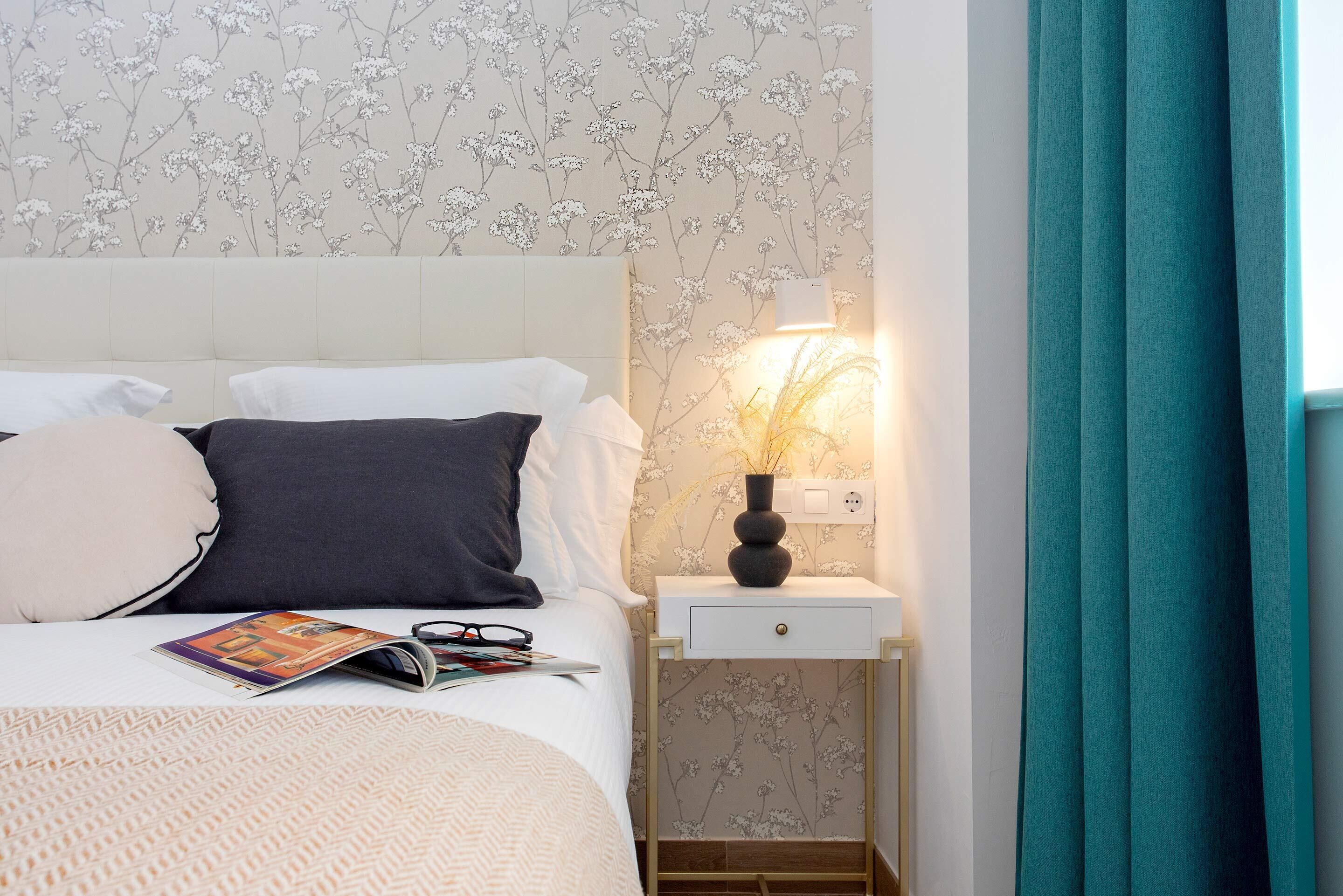 A neatly made bed with pillows, a bedside table with a lamp, and a floral wallpaper.