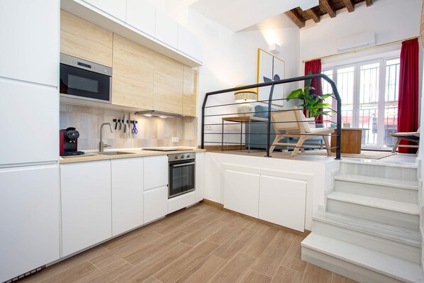 Une cuisine moderne avec des meubles blancs, un four encastré et un four à micro-ondes. Un escalier mène à un balcon agrémenté d’un rideau rouge et d’une plante en pot.