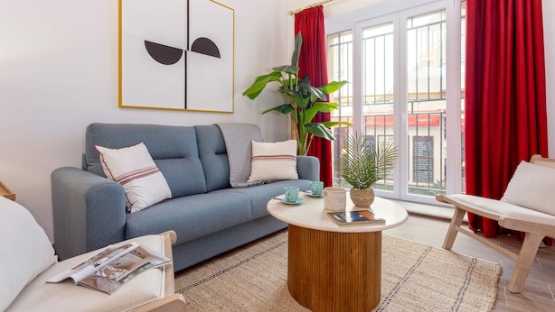A modern living room with a blue sofa, a round wooden coffee table, and a view of the outside through large windows with red curtains. - Luxury Apartment in Sevilla Center. Pajaritos V (Seville)