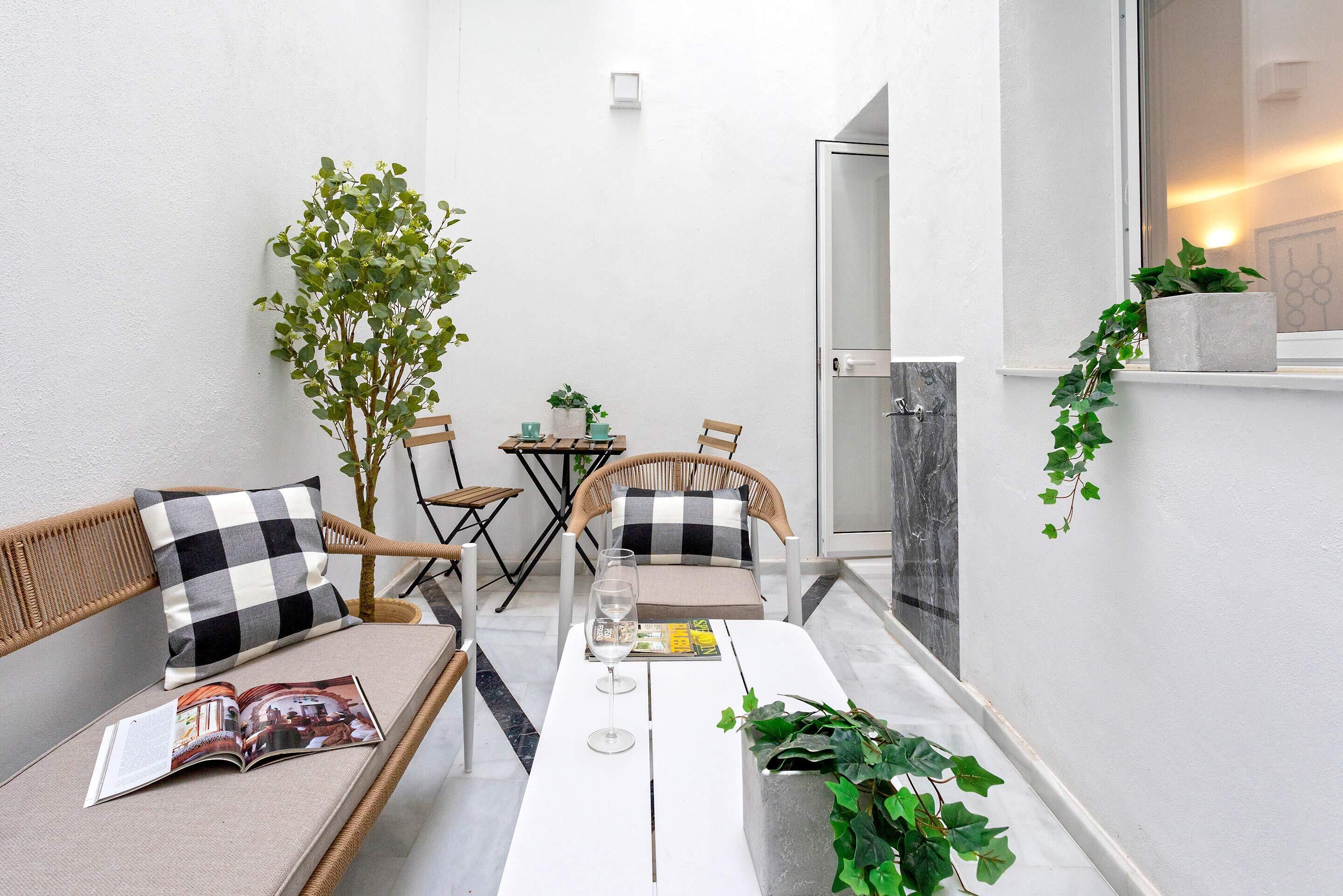 Een klein, modern buiten zitgedeelte met een witte tafel, twee rieten stoelen en een bank. Er is een glazen deur die naar een toilet leidt, en er staat een potplant aan de rechterkant.