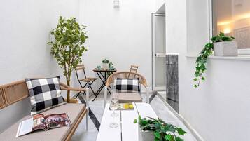 Een klein, modern buiten zitgedeelte met een witte tafel, twee rieten stoelen en een bank. Er is een glazen deur die naar een toilet leidt, en er staat een potplant aan de rechterkant.