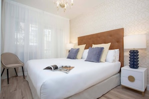 Un lit bien fait, avec des oreillers, une table de chevet avec une lampe, une chaise et une fenêtre ornée de rideaux légers.