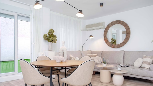 Una sala de estar moderna con una mesa de comedor redonda, sillas color beige, un sofá gris y un espejo redondo en la pared.