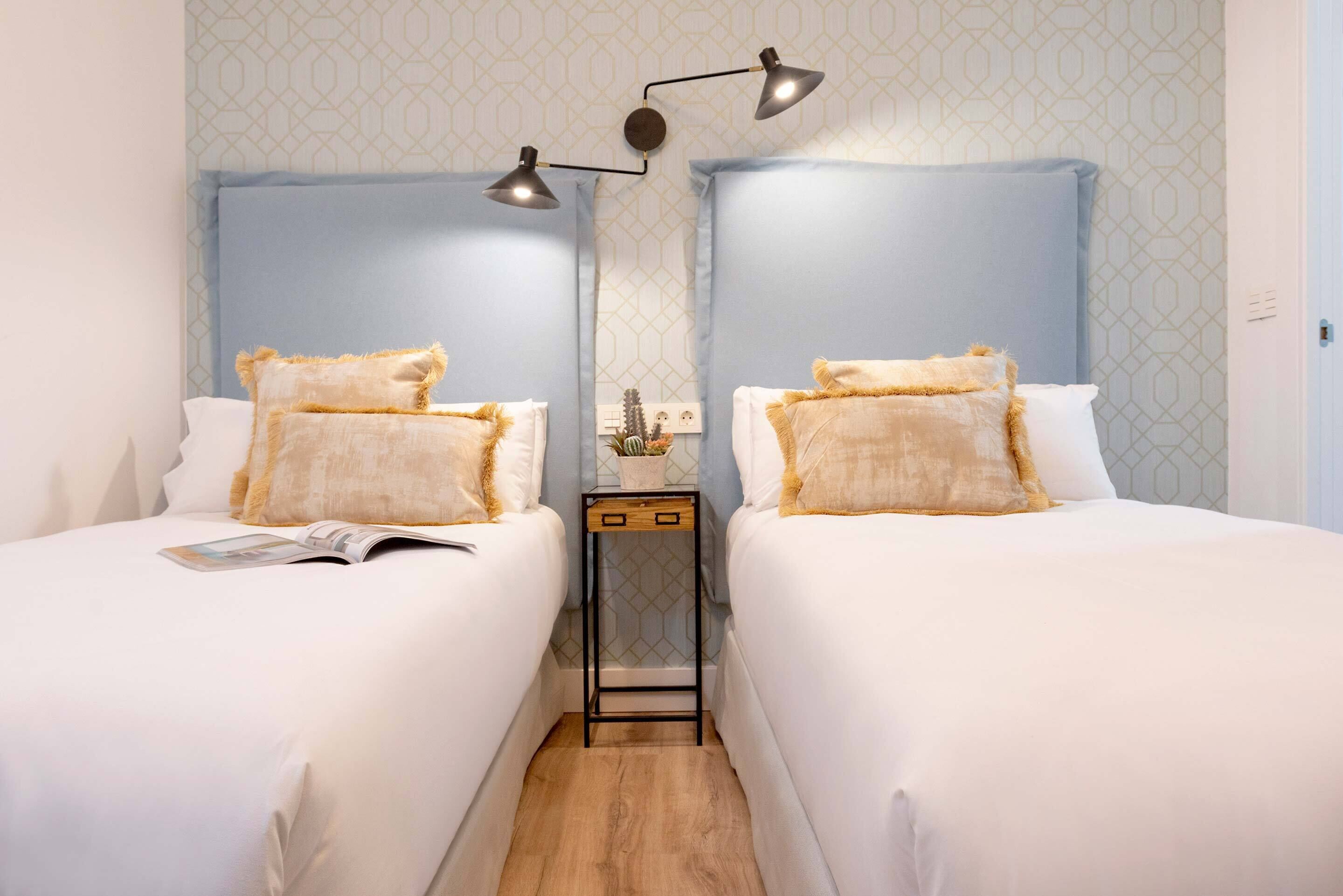 A hotel room with two beds, each with white bedding and beige pillows. There is a small table with a lamp between the beds.