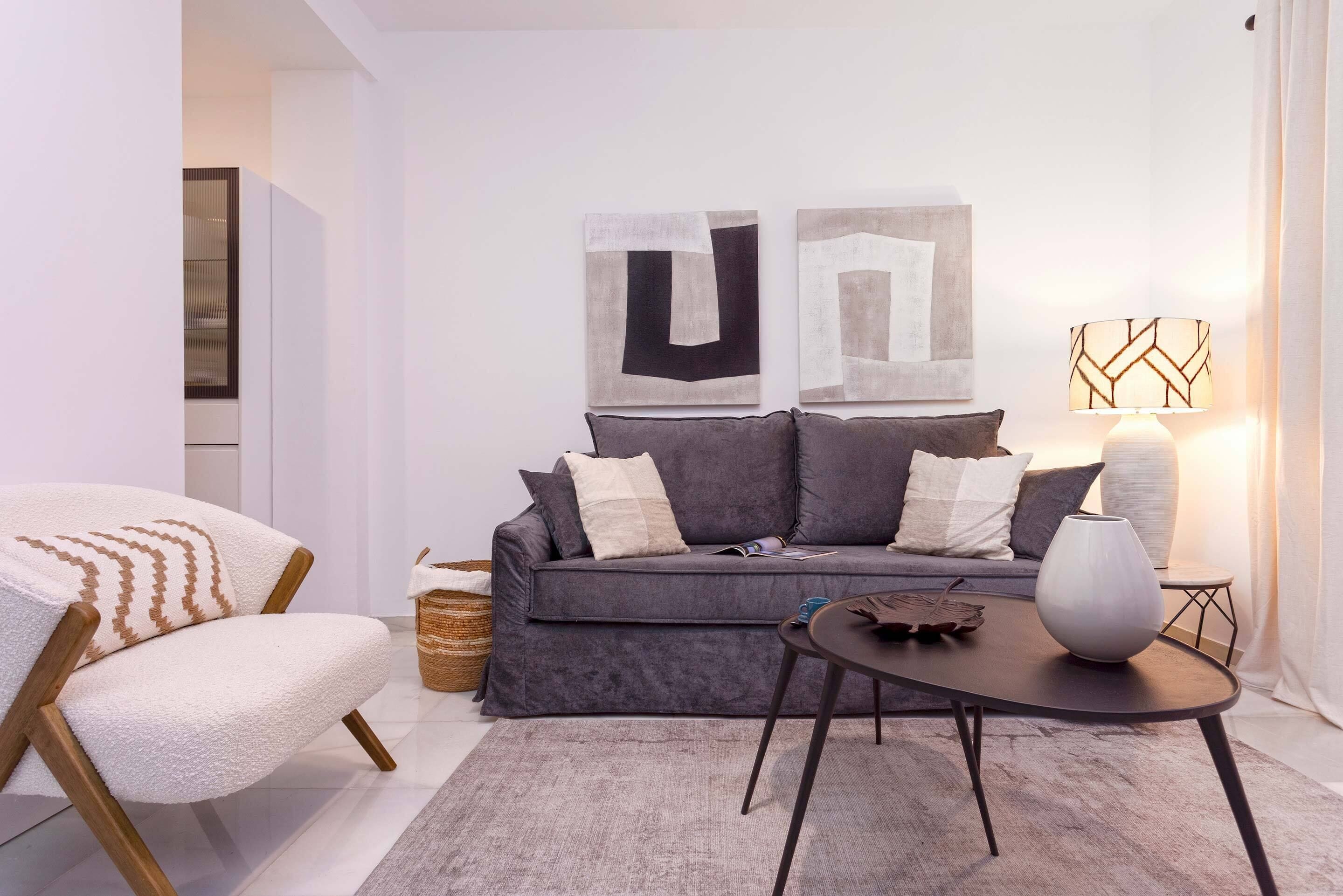 A modern living room with a grey sofa, a white armchair, and a small table with a vase. There are abstract paintings on the wall and a lamp on the table.