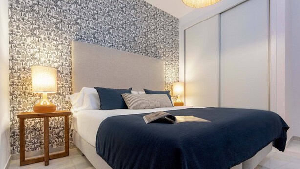 A neatly made bed with a dark blue bedspread and pillows, a wooden nightstand with a lamp, and a patterned wallpaper. - Cozy Apartment in Sevilla Center. Baños IV (Seville)