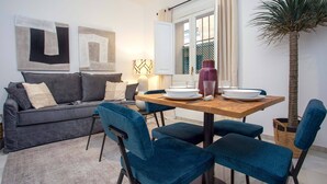A compact living space with a sofa, dining table, and chairs. There is a window with curtains, a potted plant, and abstract wall art. - Cozy Apartment in Sevilla Center. Baños IV (Seville)