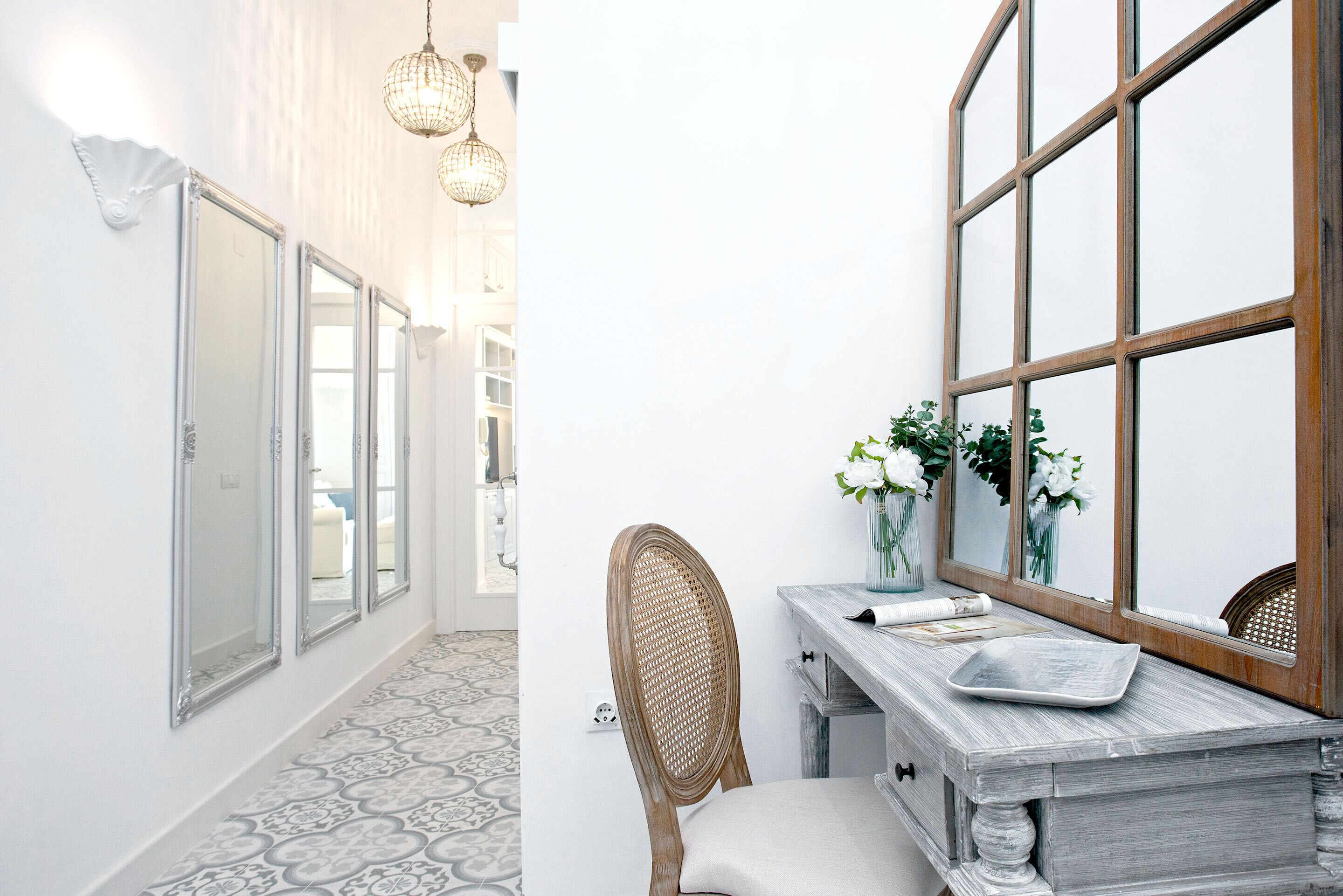 Un pasillo con suelo estampado, una silla de madera, una mesa con un jarrón de flores y un espejo con marco de madera.