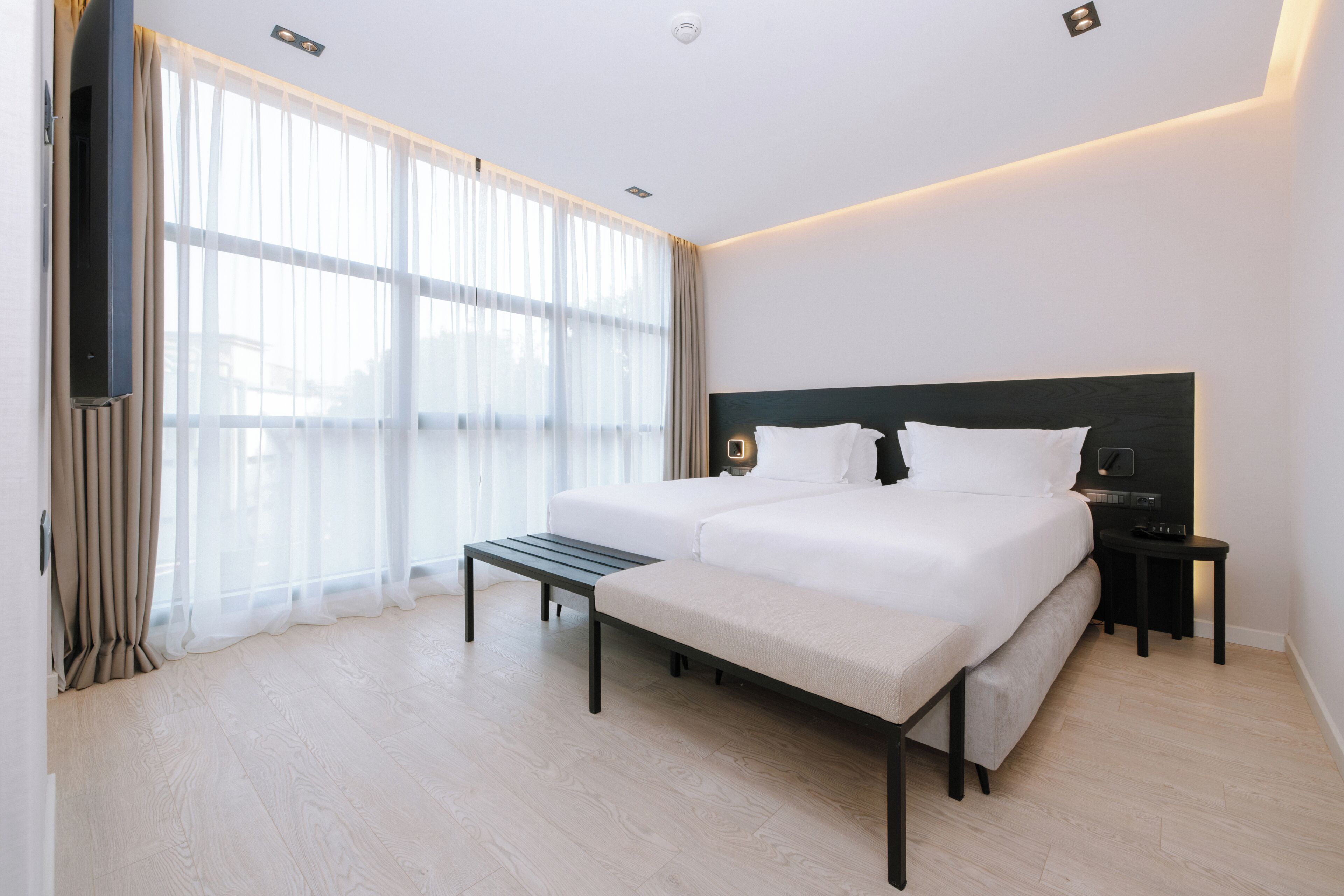 A modern hotel room with a large bed, bedside tables, a flat-screen TV, and a large window with sheer curtains.
