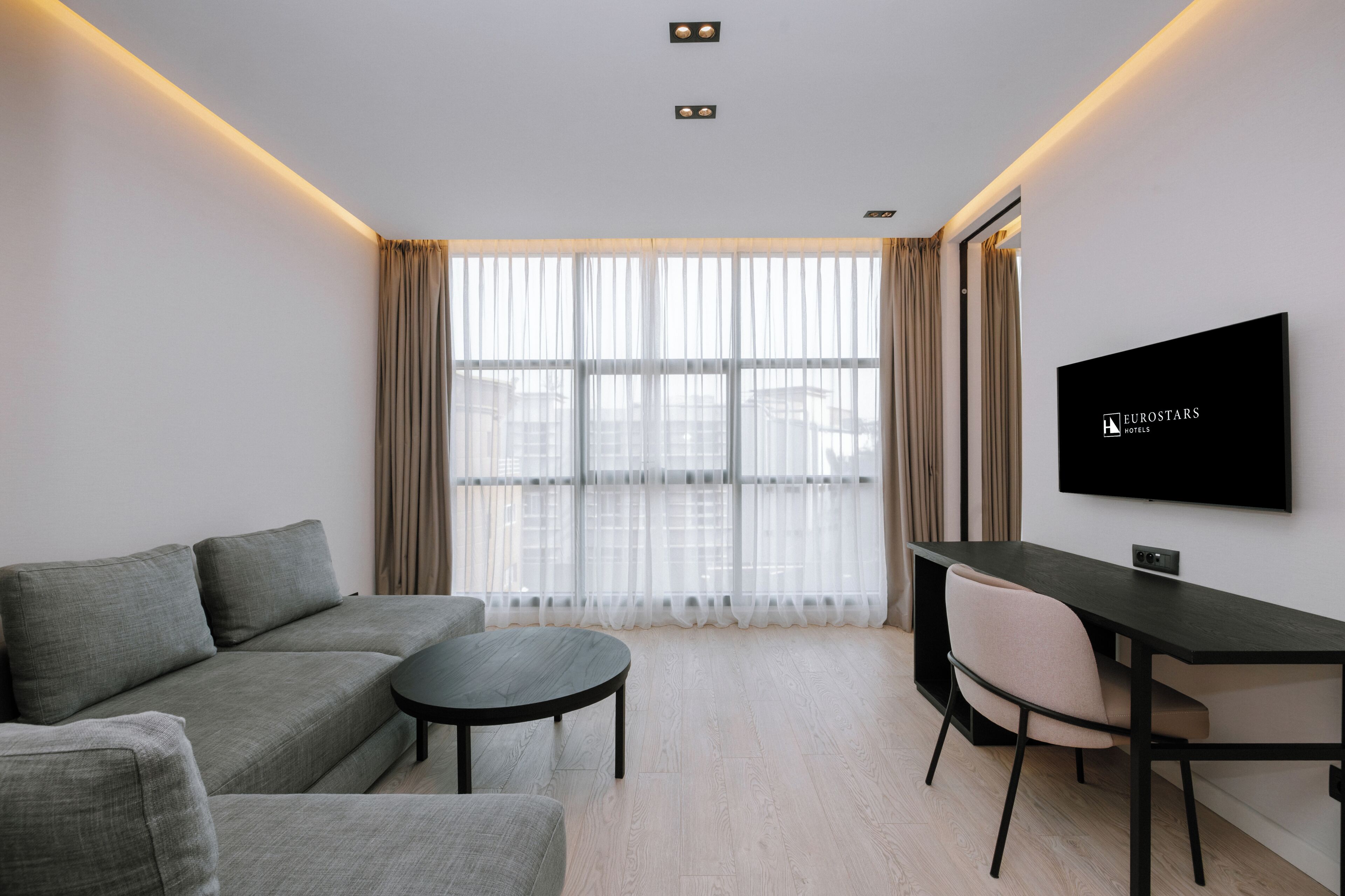 Quarto de hotel moderno com sofá, mesa redonda, televisão e grandes janelas com cortinas.
