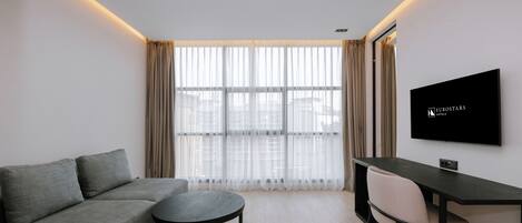 Quarto de hotel moderno com sofá, mesa redonda, televisão e grandes janelas com cortinas.