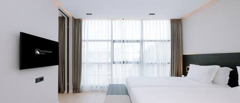 A modern hotel room with a large bed, a flat-screen TV mounted on the wall, and a large window with sheer curtains.