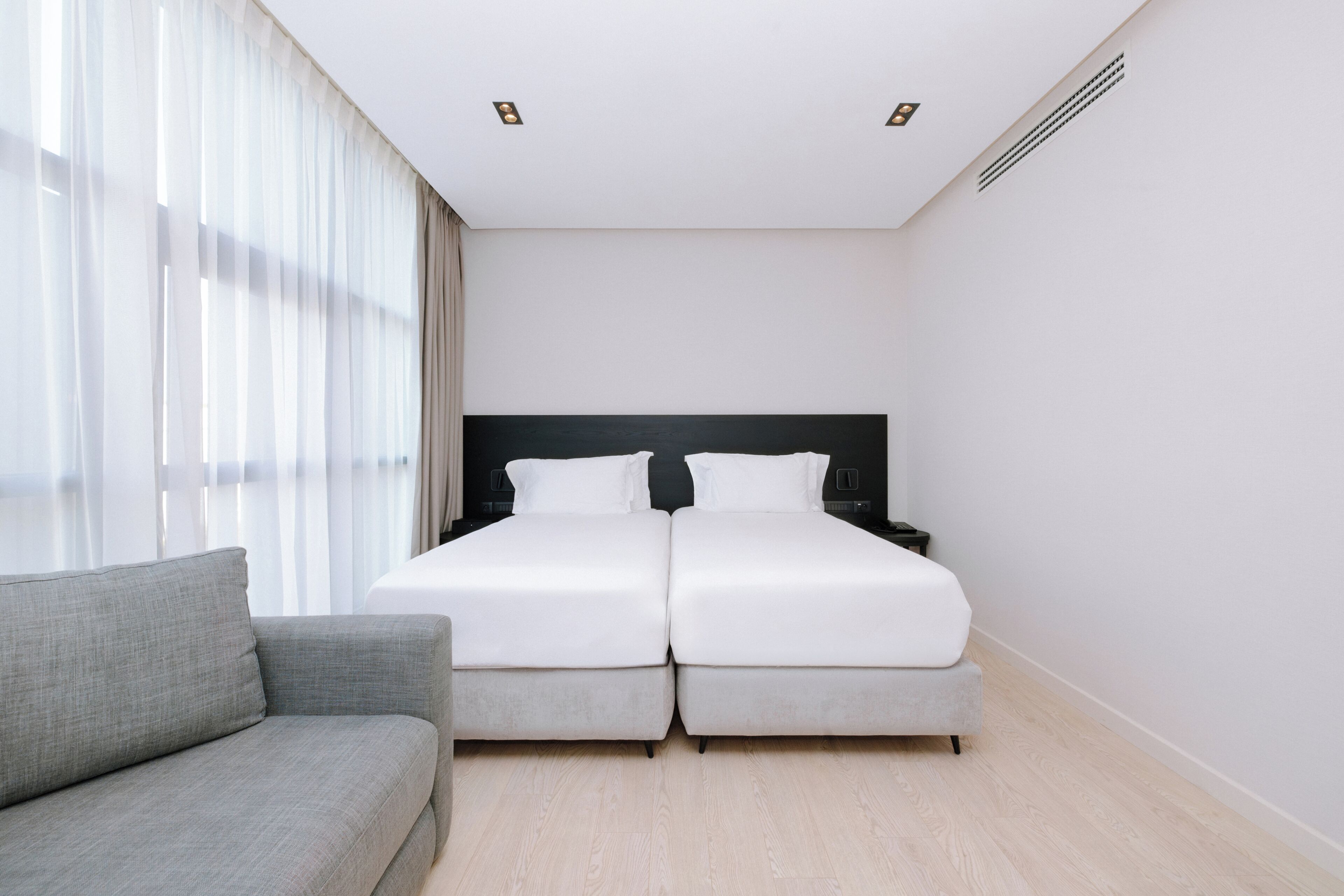 A hotel room with a double bed, a grey sofa, and a large window with sheer curtains.
