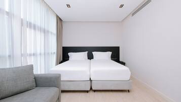 Une chambre d’hôtel avec un lit double, un canapé gris et une grande fenêtre avec des rideaux légers.