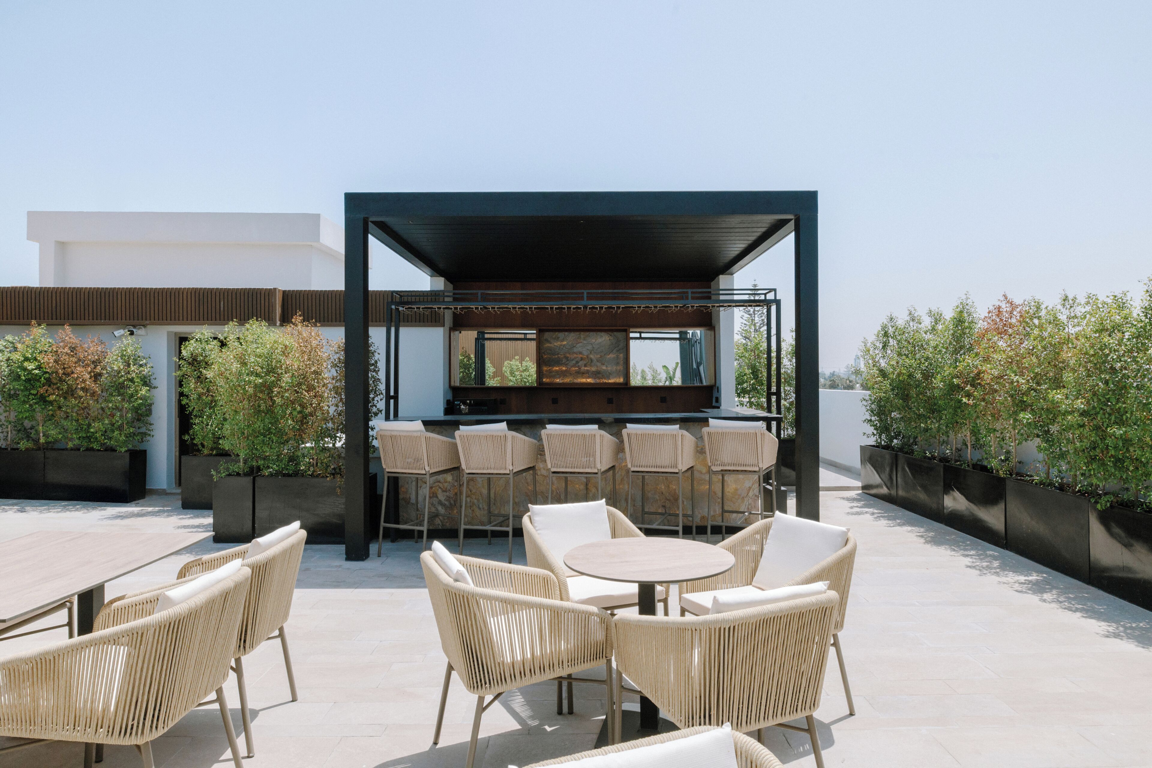 Un espacio al aire libre con sillas de mimbre y una mesa redonda, una zona de bar con una parrilla, y plantas en macetas.