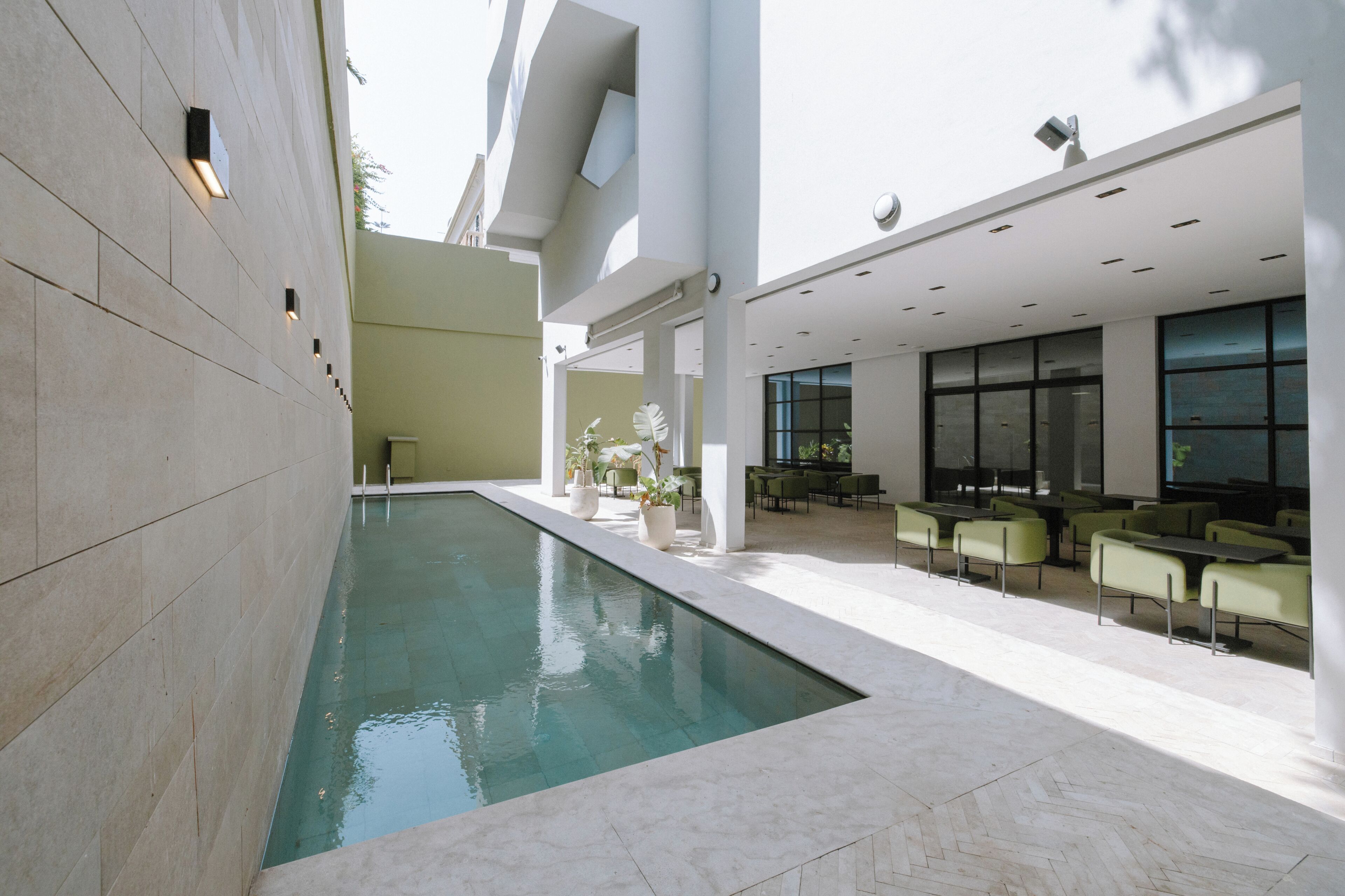 A modern indoor pool area with a clear pool, seating area with tables and chairs, and a large window providing natural light.