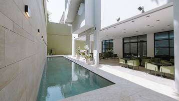 A modern indoor pool area with a clear pool, seating area with tables and chairs, and a large window providing natural light.