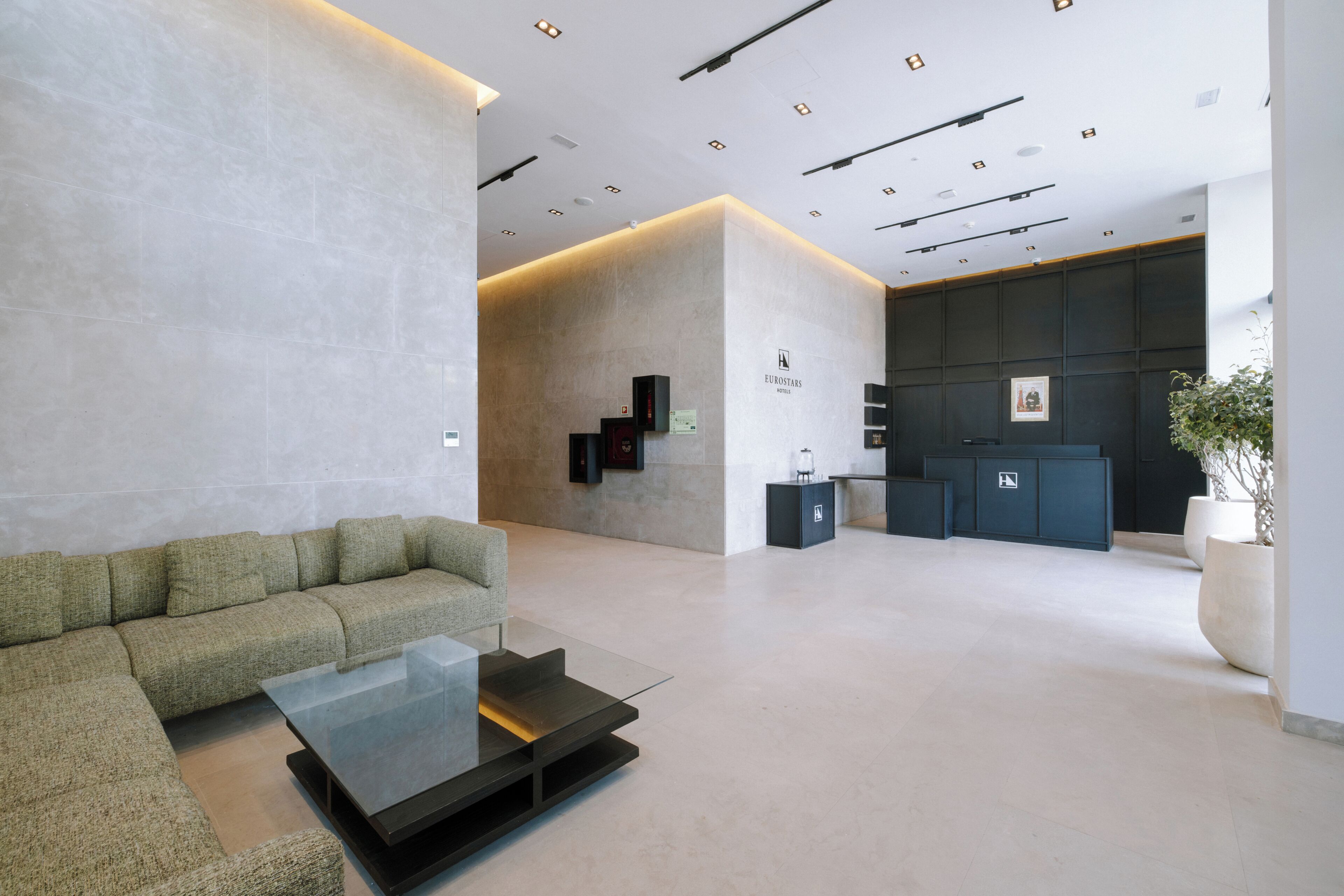 A modern lobby with a sofa, a glass coffee table, a reception desk, and a wall-mounted information board.