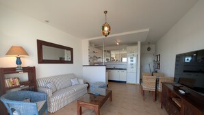 A living room with a sofa, a coffee table, a TV stand, and a kitchen area with a refrigerator and cabinets. - 182 Livingtarifa Famara (Tarifa)