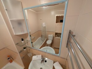 A bathroom with a bathtub, toilet, sink, and a large mirror. There are shelves with toiletries and a small plant.