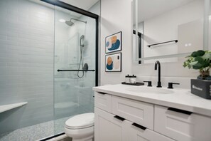 Ein modernes Badezimmer mit Duschkabine aus Glas, weiß gefliesten Wänden, einem weißen Waschbecken und einer Toilette. An der Wand befinden sich zwei gerahmte abstrakte Kunstwerke.