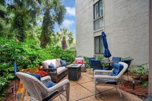 Eine Terrasse im Freien mit Korbmöbeln, einem Tisch und Stühlen, umgeben von üppiger Bepflanzung und Palmen.