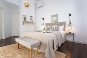 Quarto com cama, mesas de cabeceira, um banquinho, prateleira fixa com objetos decorativos e luminária de parede.