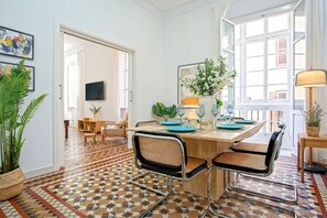 Uma sala de jantar com uma mesa de madeira, duas cadeiras de vime, um vaso com flores e uma grande janela com cortinas.