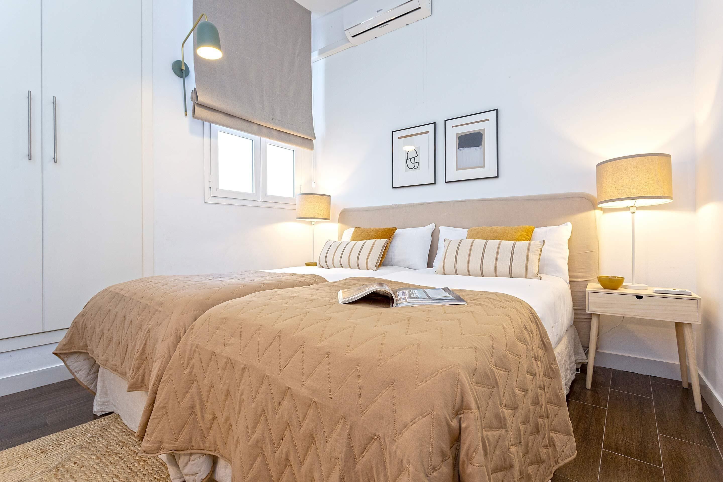 A bedroom with two beds, a bedside table with a lamp, a window with blinds, and a framed picture on the wall.