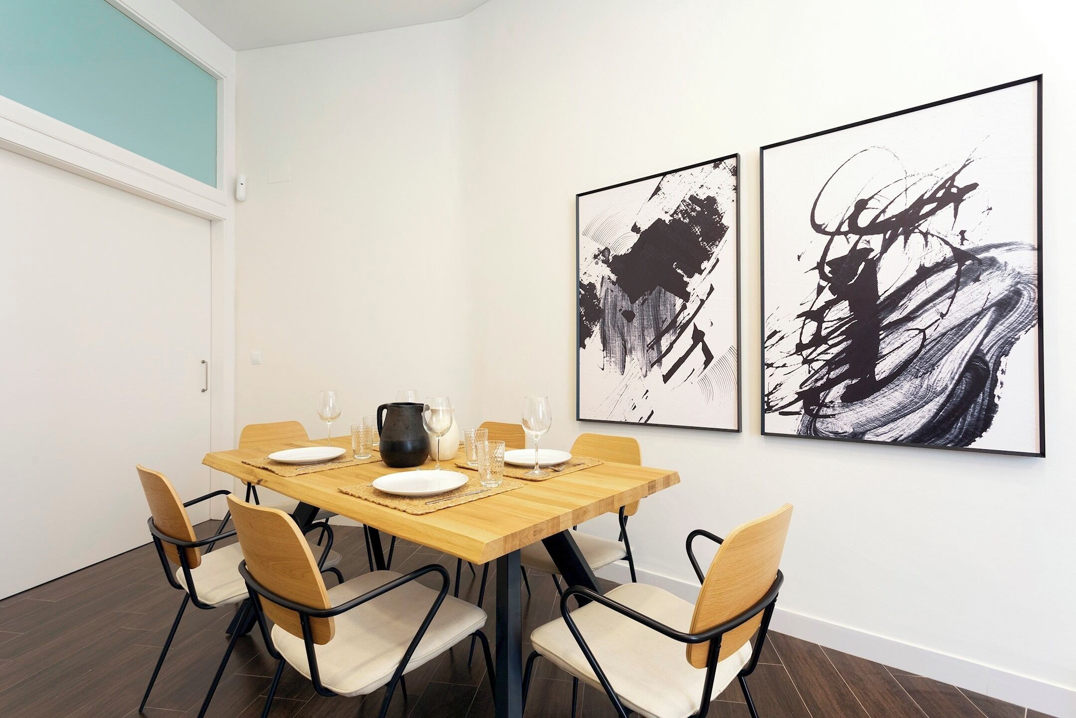 A dining area with a wooden table, four chairs, and two abstract paintings on the wall.