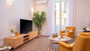 A modern living room with a flat-screen TV, a potted plant, a dining table with chairs, and a large window with curtains.