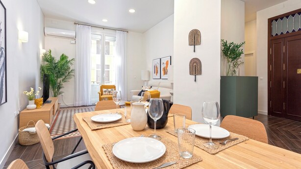 A dining area with a wooden table set for four, surrounded by chairs. There are two wall-mounted air conditioners, a large window with curtains, and a sofa in the background.