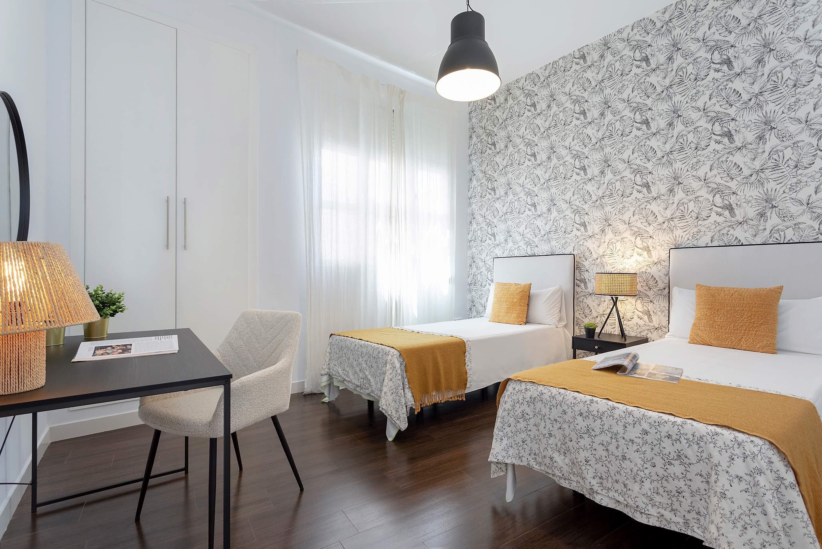 A hotel room with two beds, a desk, a chair, a lamp, and a patterned wallpaper.