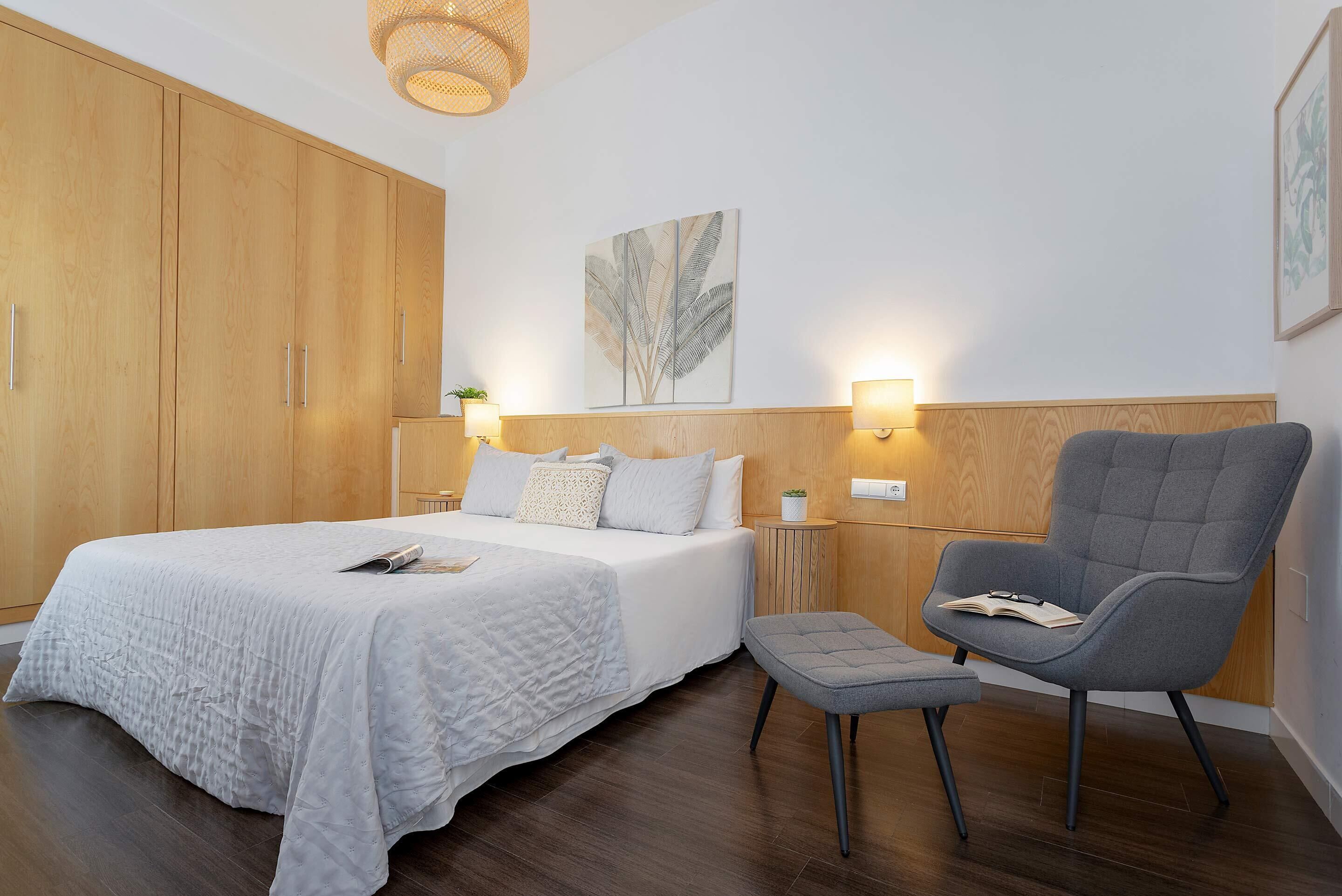 Ein Schlafzimmer mit einem großen Bett, einem grauen Sessel, einem kleinen Tisch und einer Wandlampe.