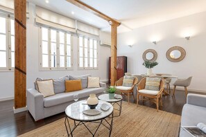 A modern living room with a grey sofa, round coffee tables, wicker chairs, and a large window with white shutters.