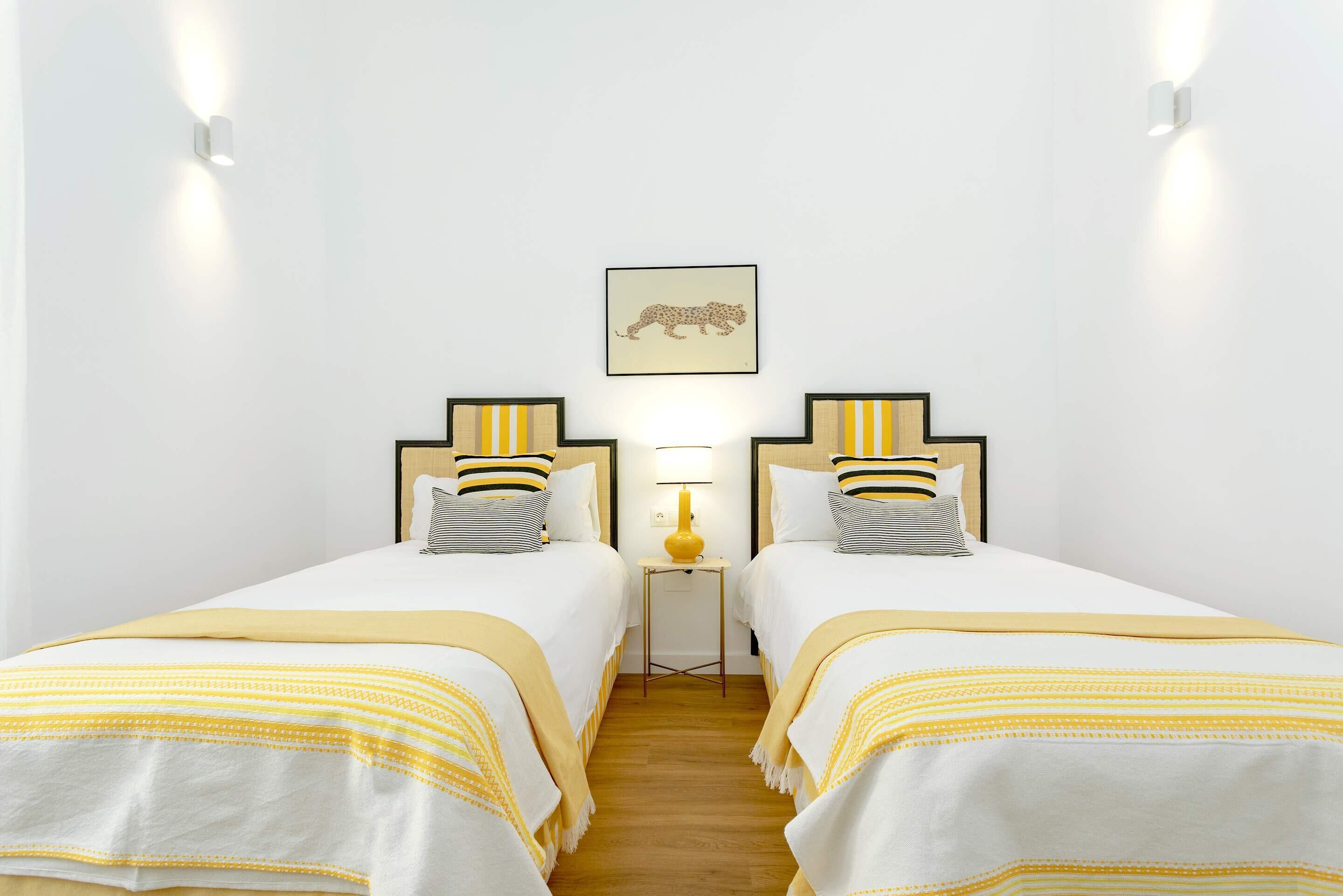 Une chambre d’hôtel avec deux lits, chacun agrémenté d’un oreiller à rayures et d’une tête de lit. Une petite table avec une lampe se trouve entre les lits, et un tableau encadré est accroché au mur au-dessus de la table.