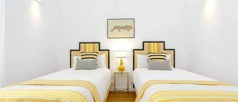 Une chambre d’hôtel avec deux lits, chacun agrémenté d’un oreiller à rayures et d’une tête de lit. Une petite table avec une lampe se trouve entre les lits, et un tableau encadré est accroché au mur au-dessus de la table.