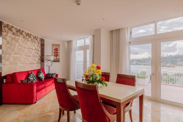 A modern living room with a red sofa, a dining table with chairs, and a balcony with a view.