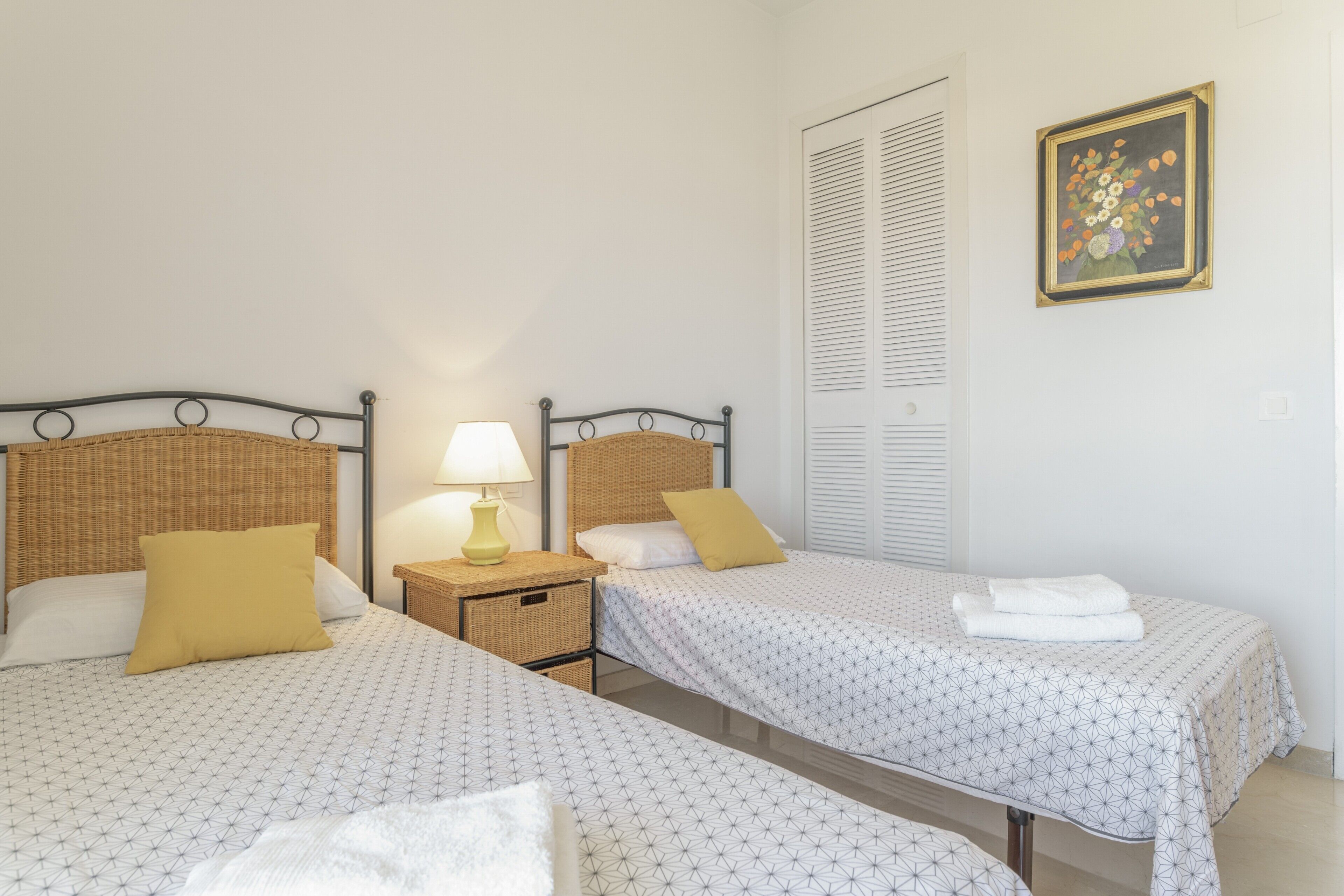 Una habitación con dos camas, una mesita de noche con lámpara, una cesta de mimbre y un cuadro enmarcado en la pared.