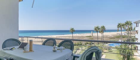 A balcony with a table and chairs, overlooking a beach and pool.