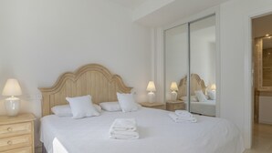 Een hotelkamer met een groot bed, twee nachtkastjes met lampen, een houten hoofdbord en een schuifdeur van glas naar een badkamer.
