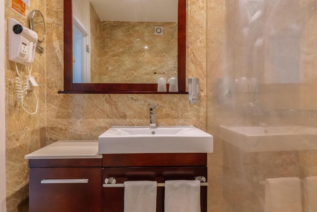 A bathroom with a marble wall, a sink, a mirror, a hairdryer, and a shower.