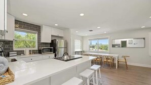 Een moderne keuken met witte aanstootbladen, een centraal eiland met krukken, een eethoek met houten stoelen en uitzicht op groen door grote ramen.