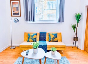 Un salon avec un canapé, deux tables d’appoint, une lampe sur pied et une fenêtre avec des rideaux.