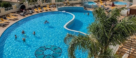 A large, curved swimming pool with multiple lanes, surrounded by palm trees and sun loungers.